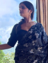 Woman wearing a black blouse with black saree with floral patterns, standing outdoors.